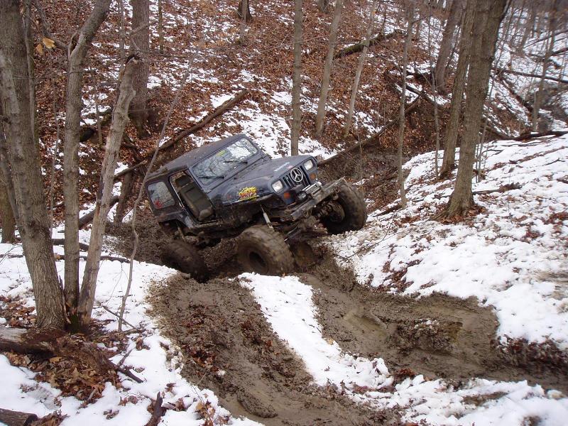 Dark Jeep Wrangler with large tires stuck in mud and snow on a wooded hill