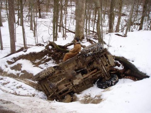 1992 Ford Bronco - An off-road vehicle lies overturned in a snow-covered ditch, with a person seated on its exposed undercarriage.