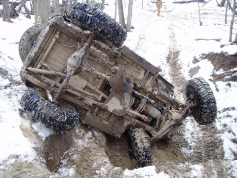 1992 Ford Bronco - An overturned off-road vehicle lies stuck in a muddy, snowy ditch, with a person walking in the background.