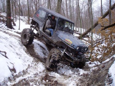 1992 Ford Bronco - A heavily modified off-road vehicle navigates a challenging snowy and muddy path through a wooded area.