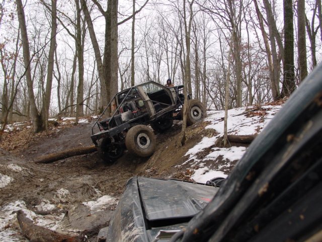 Off-road buggy with roll cage climbing a muddy, wooded hill; another vehicle in foreground