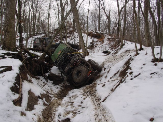 Two off-road vehicles are stuck in a snowy ditch in a wooded area. One is on its side