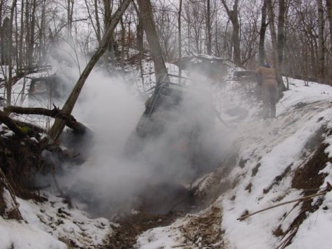 1992 Ford Bronco - An off-road vehicle is partially submerged in a snowy ditch, surrounded by trees and emitting smoke.