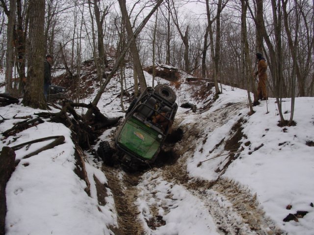 Green off-road vehicle tilted in a snowy ditch with a spare tire visible on top
