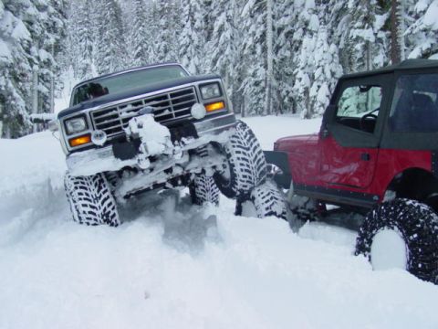 1986 Ford Bronco - Two 4x4 vehicles struggle in a snowy forest, one appears to be a Ford Bronco.