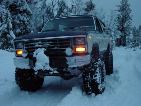 1986 Ford Bronco - A lifted Ford Bronco navigates a snow-covered trail in a wooded area.
