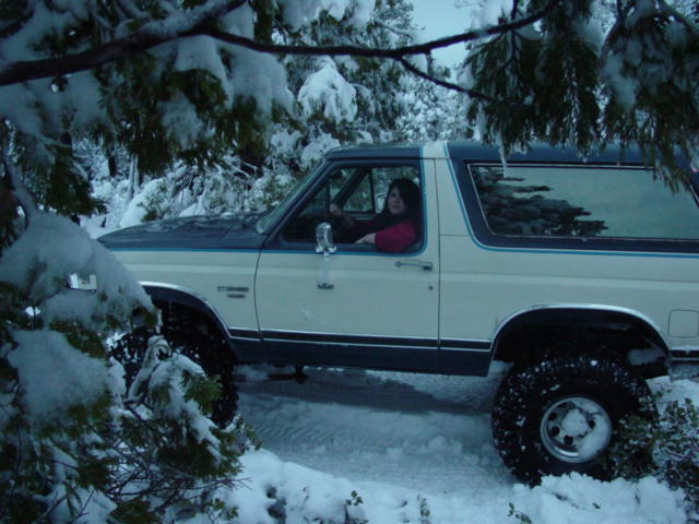 White and blue 1987 Ford Bronco in snow, side view. Tires are large and knobby. A woman is in the driver's seat