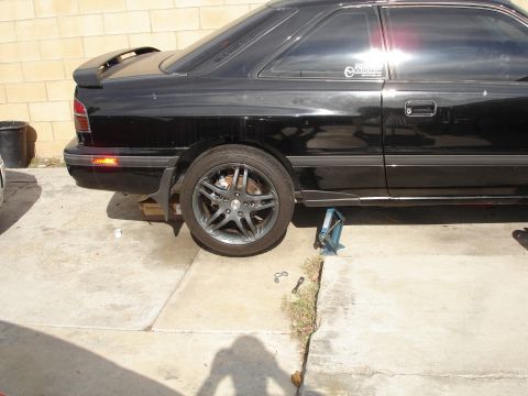 1989 Mazda MX-6 - A black coupe is shown from the side, with its rear passenger wheel removed and the car supported by a jack.
