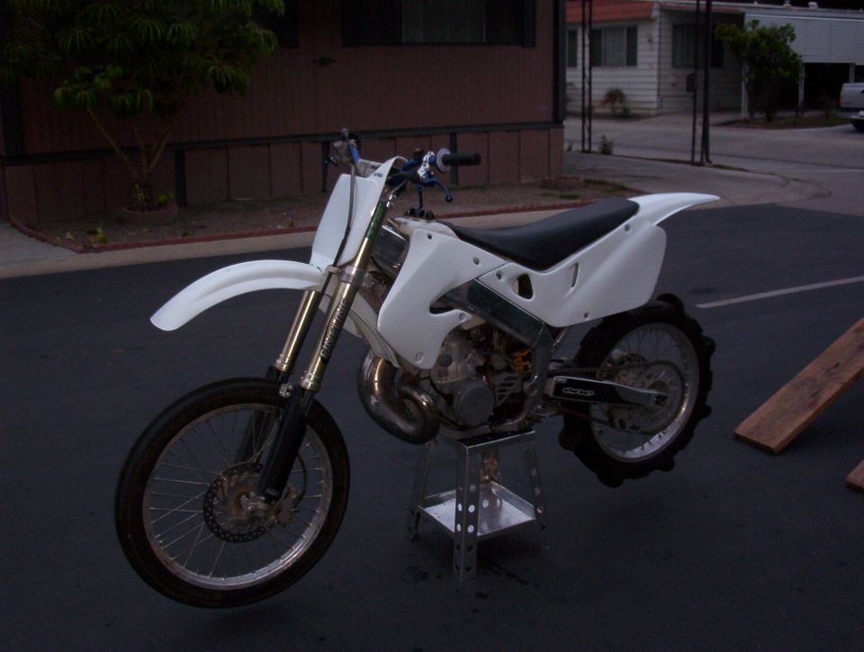 White-plastic dirt bike on a stand in a paved lot near a mobile home at dusk