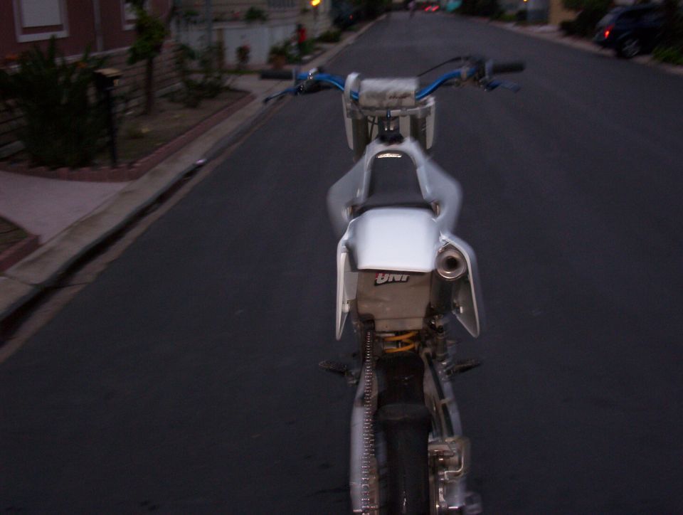 Dirt bike rear view, showing seat, exhaust, and chain. Text 'UNI' visible on a part