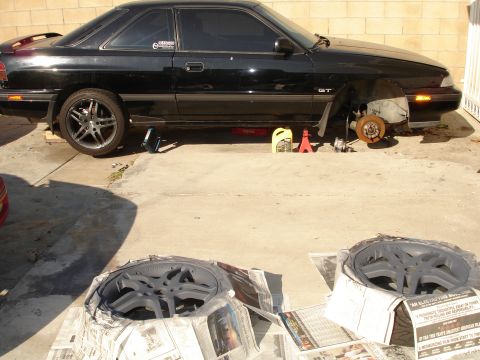 1989 Mazda MX-6 - Black coupe undergoing maintenance with wheels off and painted spares nearby.