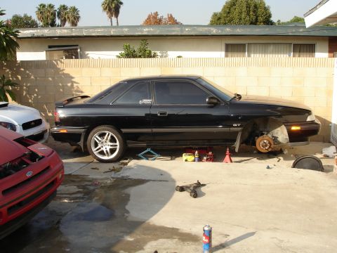 1989 Mazda MX-6 - A black 1989 Mazda MX-6 GT is parked outside with its front wheel removed, indicating ongoing maintenance or repair work.