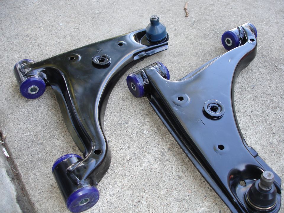 Two black control arms with blue polyurethane bushings and ball joints, resting on concrete