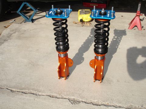 1989 Mazda MX-6 - Two orange coilover suspension assemblies with black springs and blue top mounts sit on a concrete surface.
