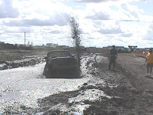 Dark pickup truck splashing through a deep mud bog pit, spraying mud high, with spectators watching