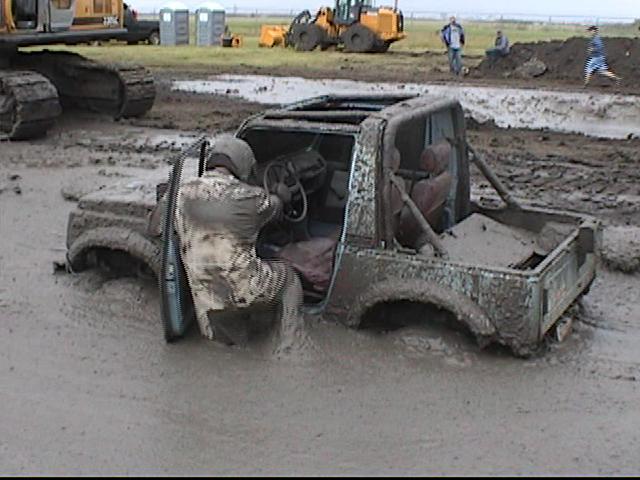 Mud-covered doorless 4x4 buried in a mud pit as the driver climbs out
