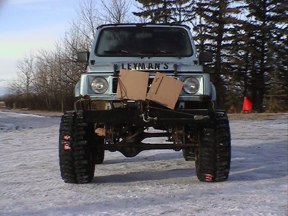 Front view of lifted light blue 4x4 with big mud tires, cardboard over grille, and 'LEYMAN'S' lettering, parked on snow