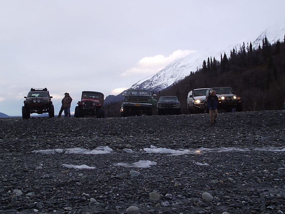 Several off-road vehicles parked on a rocky surface with a snow-capped mountain and trees in the background
