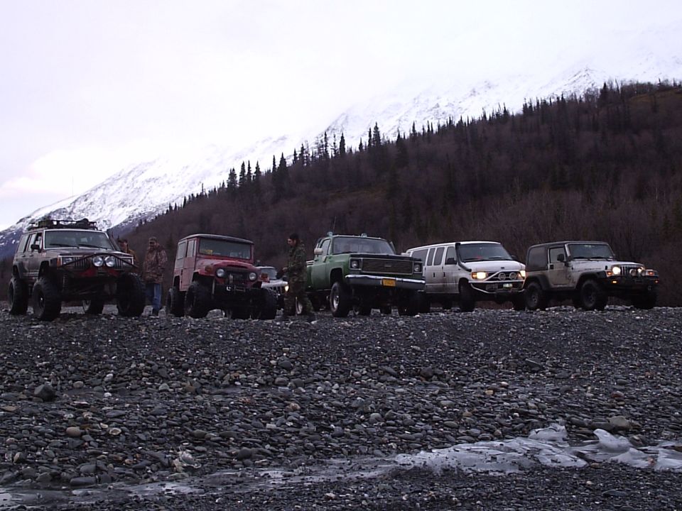 Several modified off-road vehicles parked on a rocky shore with a snow-capped mountain in the background