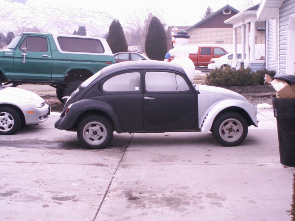 Side view of a two-tone Volkswagen Beetle, one side is black, the other is primer gray. A green Ford Bronco is parked behind it