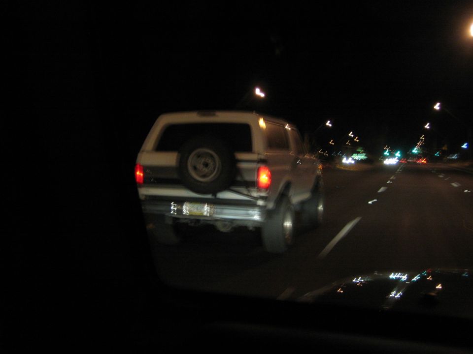 White SUV driving at night, spare tire mounted on rear door. License plate is visible but unreadable