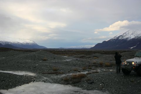 2003 Jeep TJ - A person stands next to a Jeep Wrangler in a vast, rocky terrain with snow-covered mountains under a cloudy sky.