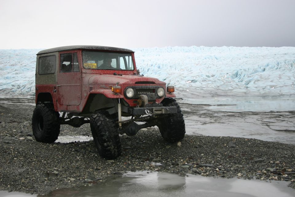 Red Toyota Land Cruiser FJ40 with large tires parked on a gravel shore in front of a glacier