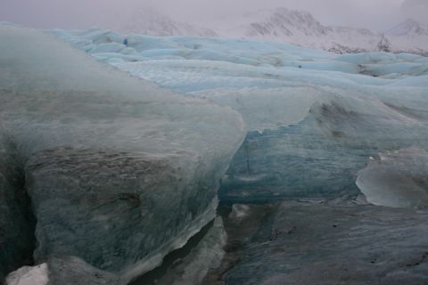 2003 Jeep TJ - The textured surface of a glacier shows layers of ice and rock under a cloudy sky with distant mountains.