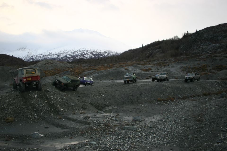 Several off-road vehicles drive on a rocky, barren landscape under a cloudy sky with snow-capped mountains