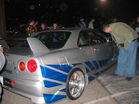 1986 Ford Bronco - A silver Nissan Skyline GT-R R34 with distinctive blue graphics is admired by a group of people at night.