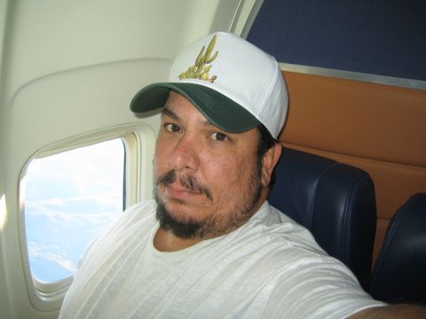 1968 Ford Mustang - Man in a white t-shirt and a baseball cap with a cactus design sits in an airplane seat, looking out the window.