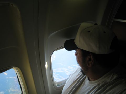 1968 Ford Mustang - A man wearing a baseball cap gazes out of an airplane window at the landscape below.