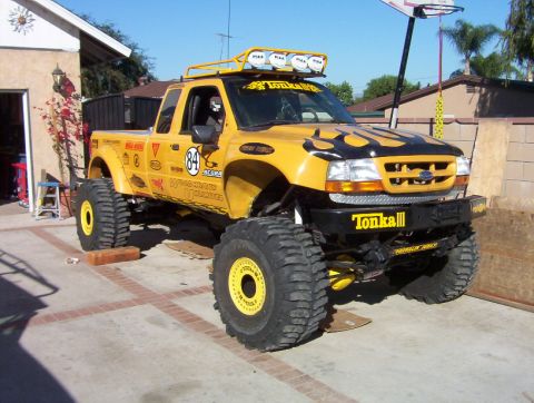 2000 Ford F-450 - Yellow and black Ford Ranger extended-cab pickup on a heavy lift with oversized bead-lock-style wheels and mud-terrain tires, parked on a d…