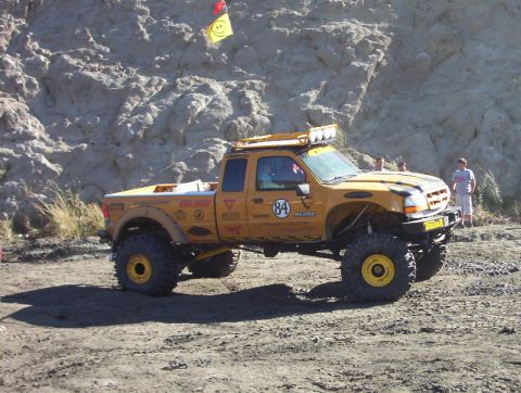 2000 Ford F-450 - Yellow extended-cab off-road pickup parked on gravel below a pale rock face, shown in profile with a yellow light bar rack over the cab, cu…