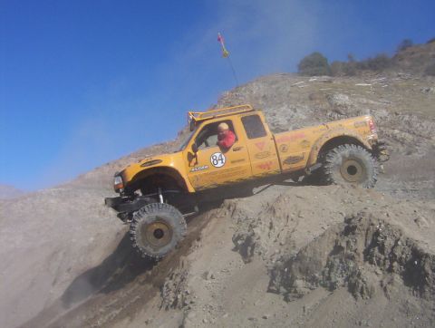 2000 Ford F-450 - A yellow lifted extended-cab pickup marked with race number 84 and sponsor decals crests a dirt berm, front wheels dropping over the edge a…