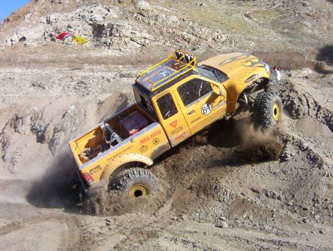 2000 Ford F-450 - Overhead angle on a yellow lifted extended-cab pickup with race number 84 door roundel descending a steep dirt bank, all four oversized tir…