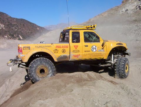 2000 Ford F-450 - Modified yellow pickup truck navigates a dirt trail with large off-road tires and a roof rack.