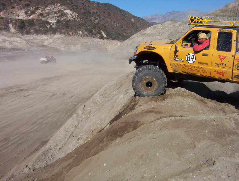 Yellow pickup truck with off-road tires and roof rack climbing a steep dirt incline. Another vehicle is visible in the background kicking up dust