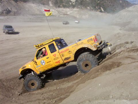 2000 Ford F-450 - A modified yellow pickup truck navigates a steep, dusty incline with other vehicles visible in the distance.
