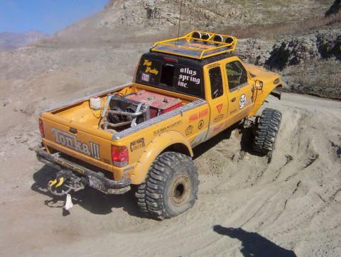 2000 Ford F-450 - High rear three-quarter view of a yellow extended-cab off-road pickup on a dirt course, its bed carrying a red generator/welder unit and a…