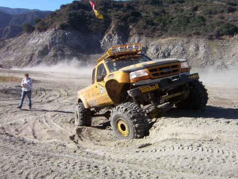 2000 Ford F-450 - Yellow Ford Ranger off-road truck flexing over a dirt mound in a dusty canyon staging area, front tire lifted and suspension articulated. I…