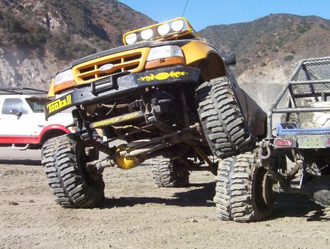 2000 Ford F-450 - A heavily lifted yellow-and-black Ford truck flexed on a dirt lot, low front angle showing the exposed solid front axle, steering linkage,…