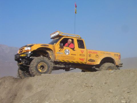 2000 Ford F-450 - Side view of a yellow extended-cab off-road pickup cresting a dirt berm against a clear blue sky, with the driver in a red shirt at the whe…