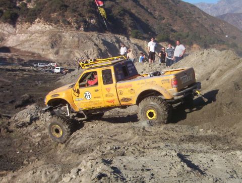 2000 Ford F-450 - Yellow extended-cab competition rock crawler flexing over a dirt hillclimb course, wearing a yellow exo/roll cage, sponsor decals, a '84' d…