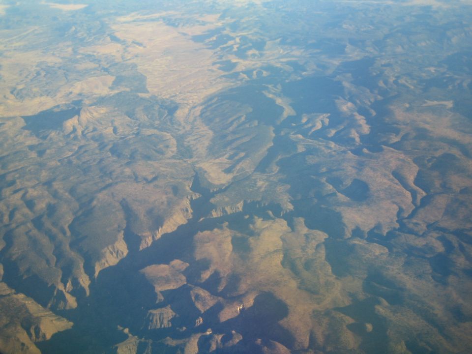 Aerial view of a vast, arid landscape with canyons and mountains under a hazy sky