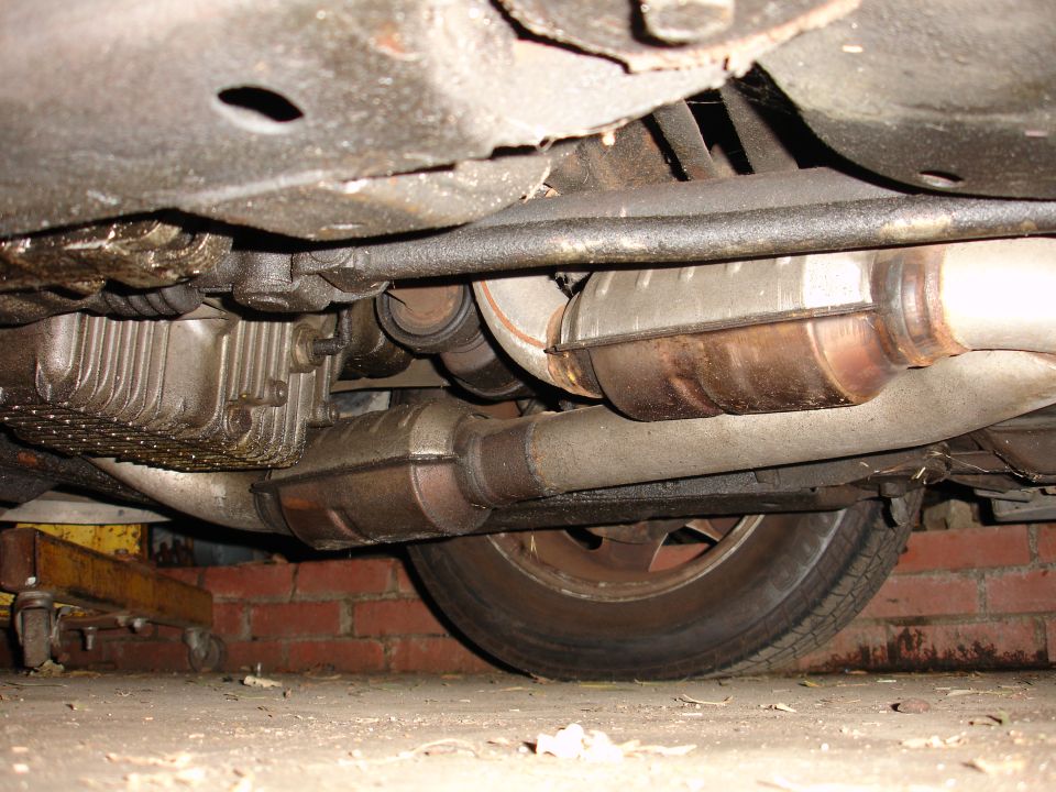 Underside view of a vehicle's exhaust system, catalytic converter, and transmission components. A wheel is visible