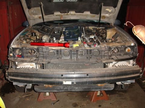1992 Ford Taurus - Disassembled car front end with hood open, revealing engine components and a red flashlight on top of the grille assembly.