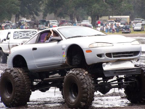 1987 Suzuki Samurai - A silver car has been heavily modified with a suspension lift and massive off-road tires, appearing in a muddy outdoor setting.