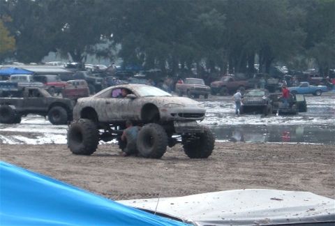 1987 Suzuki Samurai - A heavily modified car with oversized tires sits in a muddy field, surrounded by other vehicles and spectators.