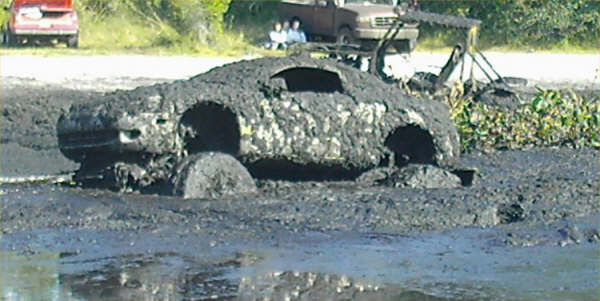 car completely covered in mud, stuck in a muddy area. another vehicle is visible in the background